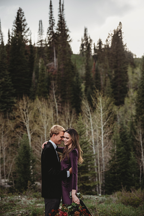 Utah-elopement-shoot-close-up-of-bride-and-groom-hugging-both-smiling-bride-in-a-deep-plum-velvet-dress-with-long-sleeves-and-lace-detailing-on-the-bodice-the-groom-is-wearing-a-black-velvet-jacket-with-tuxedo-pants-and-a-simple-black-bow-tie