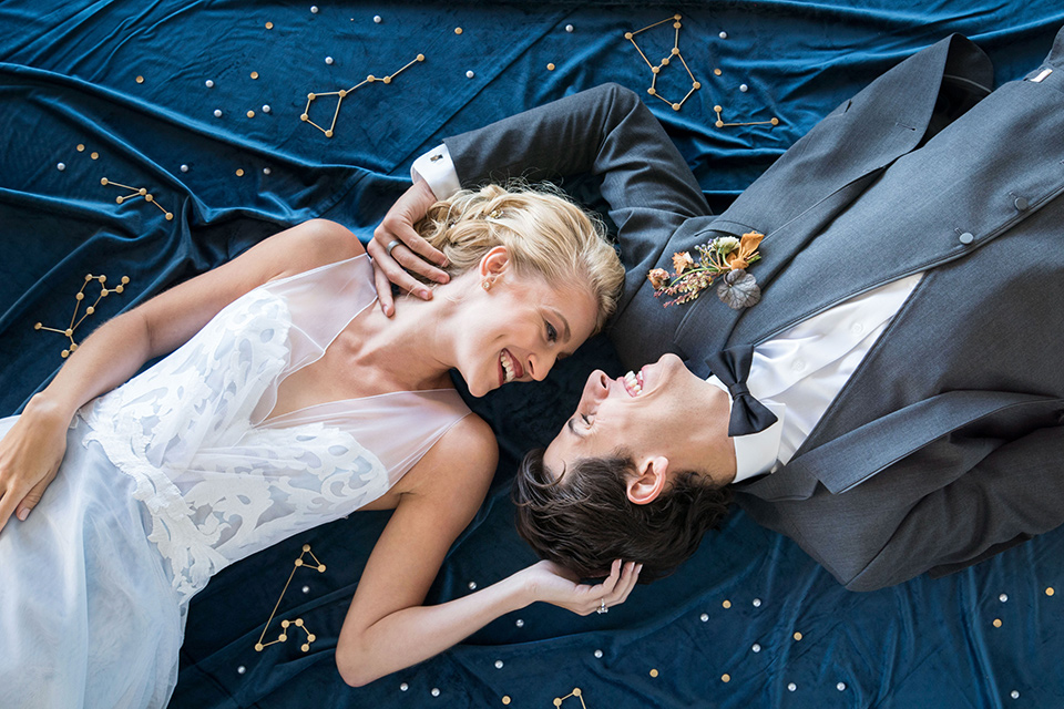 hangar-21-bride-and-groom-laying-on-blue-fabric-bride-wearing-a-blue-dress-with-lace-details-on-the-bodice-and-flowing-light-blue-material-for-the-skirt-grooom-in-a-charcoal-tuxedo-with-a-matching-charcoal-tie