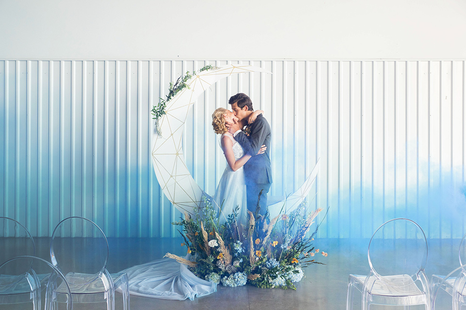 hangar-21-ceremony-brode-and-groom-kissing-with-smoke-bomb-behind-them-a-blue-dress-with-lace-details-on-the-bodice-and-flowing-light-blue-material-for-the-skirt-grooom-in-a-charcoal-tuxedo-with-a-matching-charcoal-tie