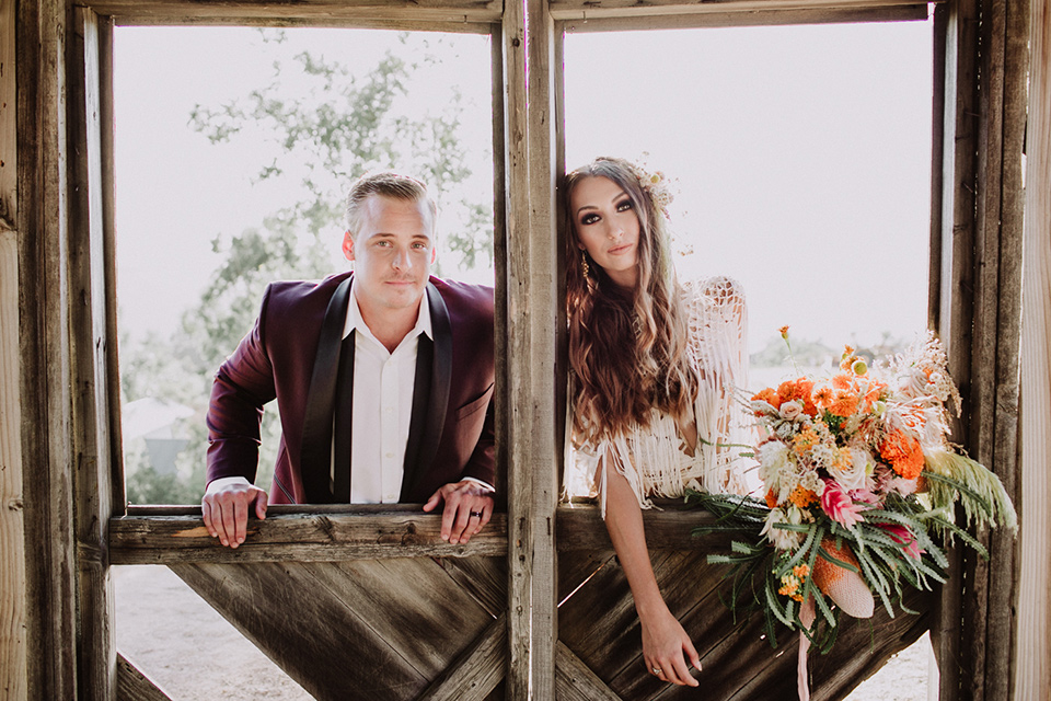 hey-babe-ranch-bride-and-groom-in-stables-bride-in-a-bohemian-gown-with-lace-and-fringe-detailing-and-hair-in-a-loose-wave-groom-in-a-burgundy-tuxedo-with-black-satin-truim-and-his-tie-undone-for-a-relaxed-look