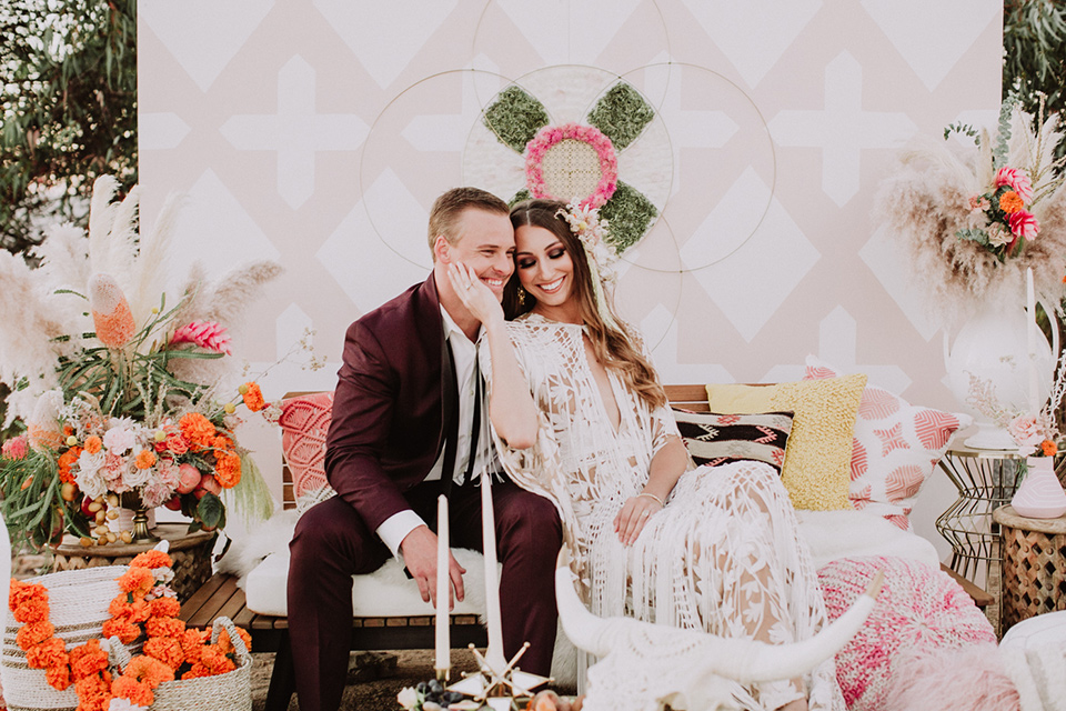 hey-babe-ranch-bride-and-groom-sitting-on-chair-laughing-bride-in-a-bohemian-gown-with-lace-and-fringe-detailing-and-hair-in-a-loose-wave-groom-in-a-burgundy-tuxedo-with-black-satin-truim-and-his-tie-undone-for-a-relaxed-look