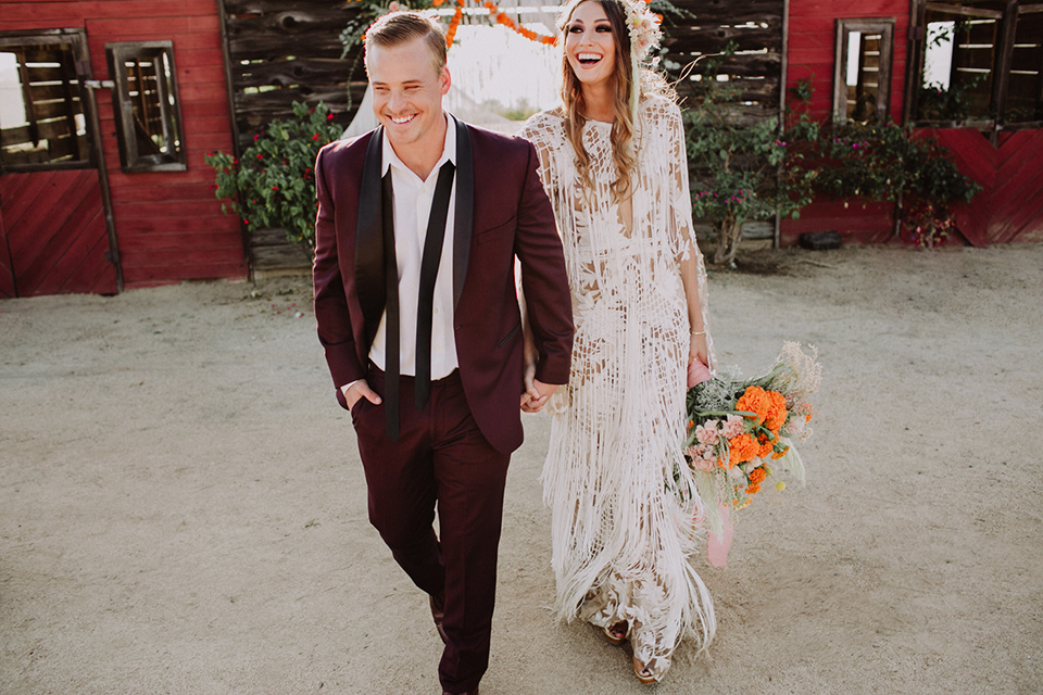 hey-babe-ranch-bride-and-groom-walking-away-and-laughing-bride-in-a-bohemian-gown-with-lace-and-fringe-detailing-and-hair-in-a-loose-wave-groom-in-a-burgundy-tuxedo-with-black-satin-truim-and-his-tie-undone-for-a-relaxed-look