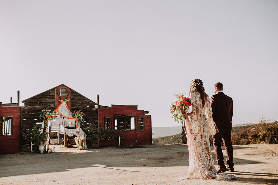 hey-babe-ranch-bride-and-groom-walking-towards-barn-bride-in-a-bohemian-gown-with-lace-and-fringe-detailing-and-hair-in-a-loose-wave-groom-in-a-burgundy-tuxedo-with-black-satin-truim-and-his-tie-undone-for-a-relaxed-look