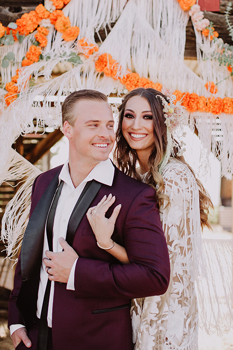 hey-babe-ranch-bride-andgroom-embracing-bride-behind-him-both-smiling-bride-in-a-bohemian-gown-with-lace-and-fringe-detailing-and-hair-in-a-loose-wave-groom-in-a-burgundy-tuxedo-with-black-satin-truim-and-his-tie-undone-for-a-relaxed-look