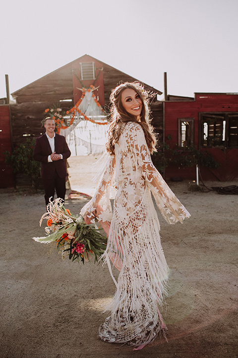 hey-babe-ranch-bride-twirling-groom-behind-her-bride-looking-at-camera-bride-in-a-bohemian-gown-with-lace-and-fringe-detailing-and-hair-in-a-loose-wave-groom-in-a-burgundy-tuxedo-with-black-satin-truim-and-his-tie-undone-for-a-relaxed-look