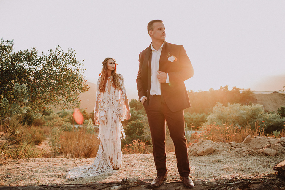 hey-babe-ranch-brode-and-groom-groom-in-foreground-looking-off-to-the-distance-hand-on-jacket-bride-in-a-bohemian-gown-with-lace-and-fringe-detailing-and-hair-in-a-loose-wave-groom-in-a-burgundy-tuxedo-with-black-satin-truim-and-his-tie-undone-for-a-relaxed-look