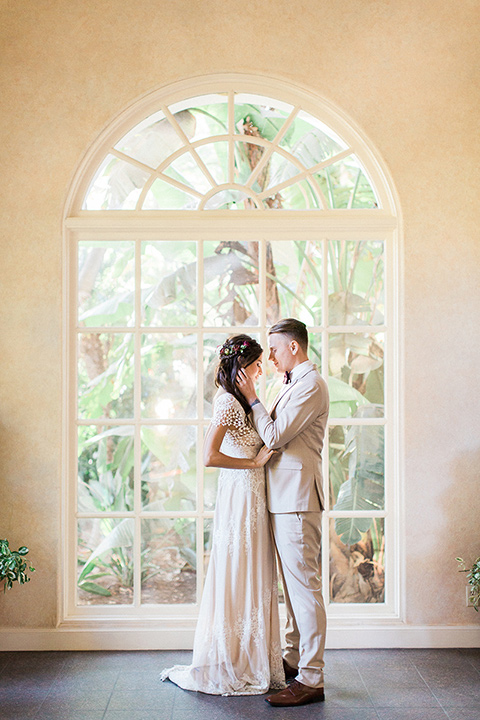 Huntington-bay-club-wedding-bride-and-groom-in-front-of-window-bride-in-a-spanish-inspired-style-dress-with-lace-detailing-and-cap-sleeves-groom-in-a-tan-suit-with-a-burgundy-matte-bow-tie-and-brown-shoes
