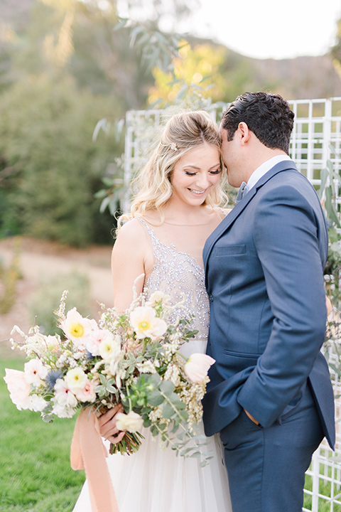 The-Lodge-at-Malibu-Lake-bride-and-groom-embrace-bride-in-a-full-ball-gown-with-a-deep-v-crystal-detailed-bodice-groom-in-a-slate-blue-suit-with-a-grey-velvet-bow-tie