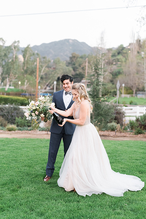 The-Lodge-at-Malibu-Lake-bride-and-groom-opeing-champs-bride-in-a-full-ball-gown-with-a-deep-v-crystal-detailed-bodice-groom-in-a-slate-blue-suit-with-a-grey-velvet-bow-tie