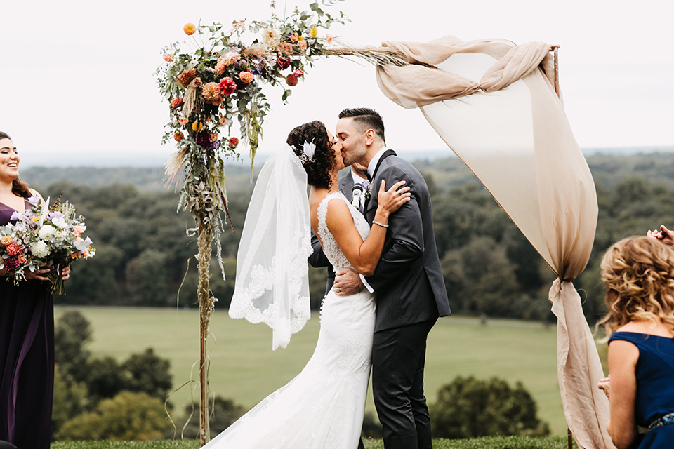 The-Mansion-at-Natirar-bride-and-grom-kiss-at-ceremony-bride-in-a-lace-fitted-dress-with-a-high-neckline-and-hair-up-groom-in-a-grey-tuxedo-with-black-trim-and-a-black-bowtie