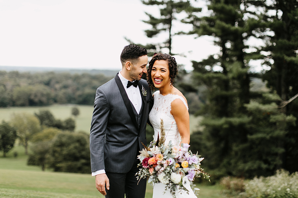 The-Mansion-at-Natirar-bride-and-groom-laughing-bride-in-a-lace-fitted-dress-with-a-high-neckline-and-hair-up-groom-in-a-grey-tuxedo-with-black-trim-and-a-black-bowtie