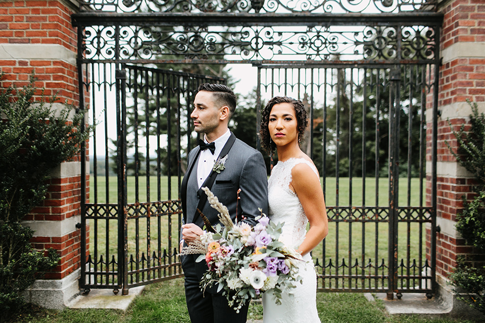 The-Mansion-at-Natirar-bride-and-groom-looking-serious-by-iron-gate-bride-in-a-lace-fitted-dress-with-a-high-neckline-and-hair-up-groom-in-a-grey-tuxedo-with-black-trim-and-a-black-bowtie