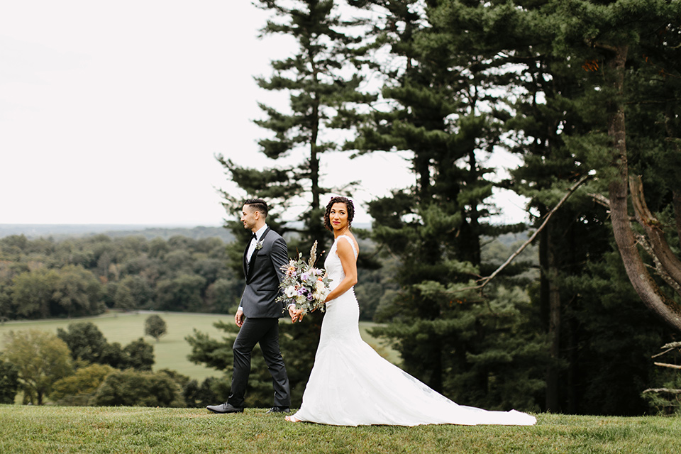 The-Mansion-at-Natirar-bride-and-groom-walking-by-the-forrest-bride-in-a-lace-fitted-dress-with-a-high-neckline-and-hair-up-groom-in-a-grey-tuxedo-with-black-trim-and-a-black-bowtie