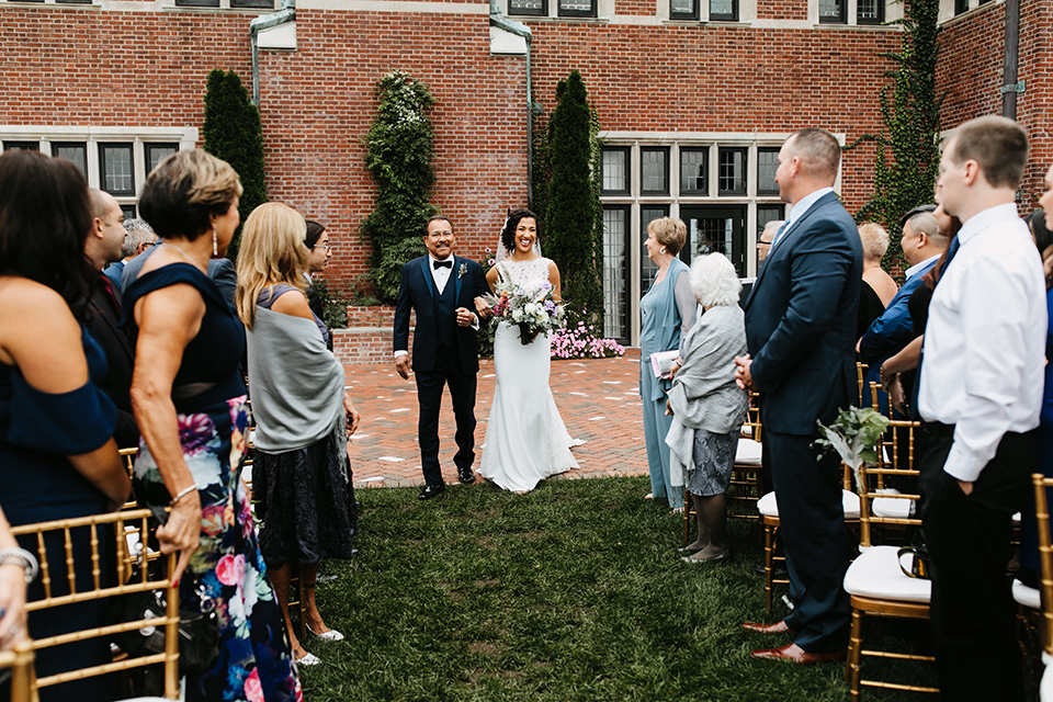 The-Mansion-at-Natirar-bride-coming-down-the-aisle-bride-in-a-lace-fitted-dress-with-a-high-neckline-and-hair-up-groom-in-a-grey-tuxedo-with-black-trim-and-a-black-bowtie