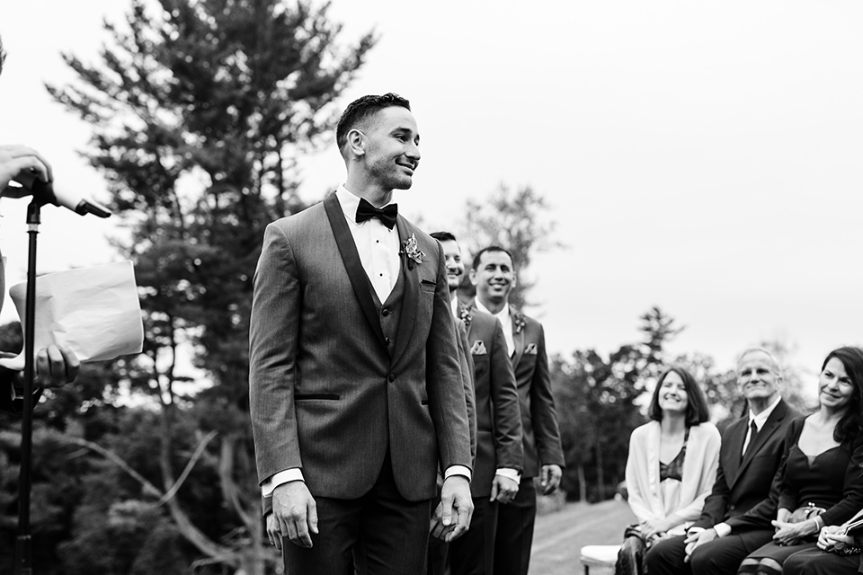 The-Mansion-at-Natirar-groom-looking-at-bride-coming-down-the-aisle-groom-in-a-grey-tuxedo-with-black-trim-and-a-black-bowtie