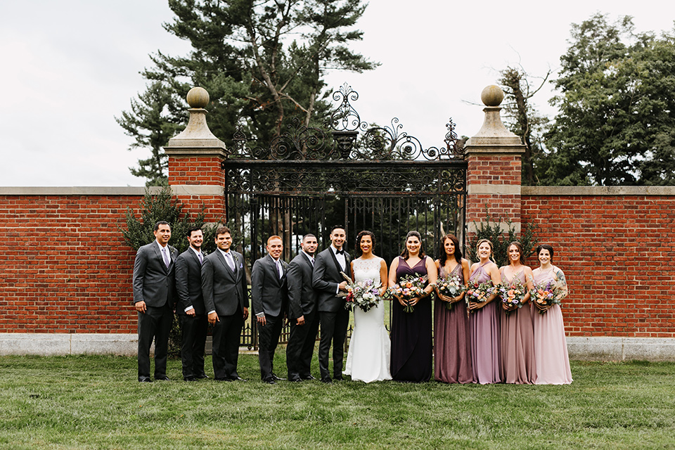 The-Mansion-at-Natirar-bridal-party-bridesmaids-in-different-shades-of-purple-dresses-groomsmen-in-grey-tuxeos-with-purple-ties-bride-in-a-lace-fitted-dress-with-a-high-neckline-and-hair-up-groom-in-a-grey-tuxedo-with-black-trim-and-a-black-bowtie