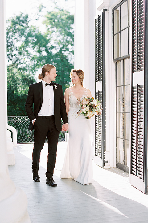 Taylor-Grady-House-shoot-bride-and-groom-walking-looking-at-each-other-bride-in-a-fit-and-flar-silk-gown-with-an-open-back-detail-and-hair-in-a-loose-bun-groom-wearing-a-black-notch-lapel-tuxedo-with-a-black-bowtie-and-hair-in-a-bun