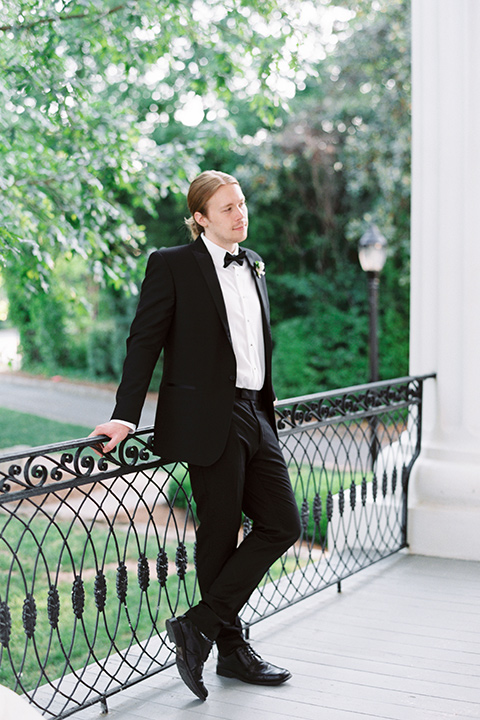 Taylor-Grady-House-shoot-groom-with-legs-crossed-groom-wearing-a-black-notch-lapel-tuxedo-with-a-black-bowtie-and-hair-in-a-bun