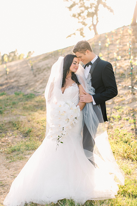 callaway-winery-shoot-groom-standing-behind-bride-in-the-vinyard-bride-wearing-a-fitted-dress-with-straps-and-tulle-and-lace-detailing-while-the-groom-wore-a-traditional-black-tuxedo-with a-black-bowtie