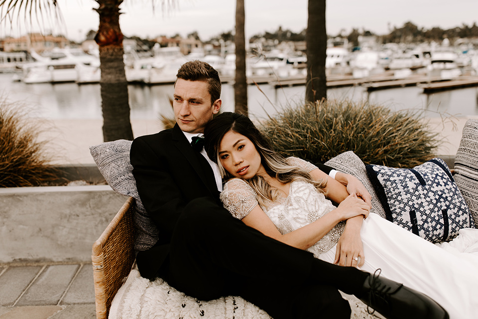 huntington-bay-club-shoot-bride-and-groo-laying-on-the-couch-bride-wearing-a-flowing-gown-with-a-beaded-bodice-and-ca-sleeves-groom-wearing-a-black-suit-with-a-green-velvet-bow-tie