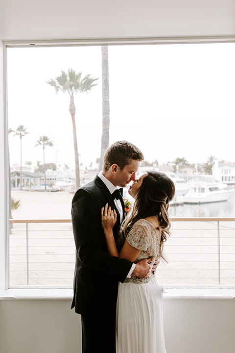 huntington-bay-club-shoot-bride-and-groom-by-window-close-up-bride-wearing-a-flowing-dress-with-a-beaded-bodice-and-sleeves-groom-wearing-a-black-suit-with-a-green-velvet-bow-tie