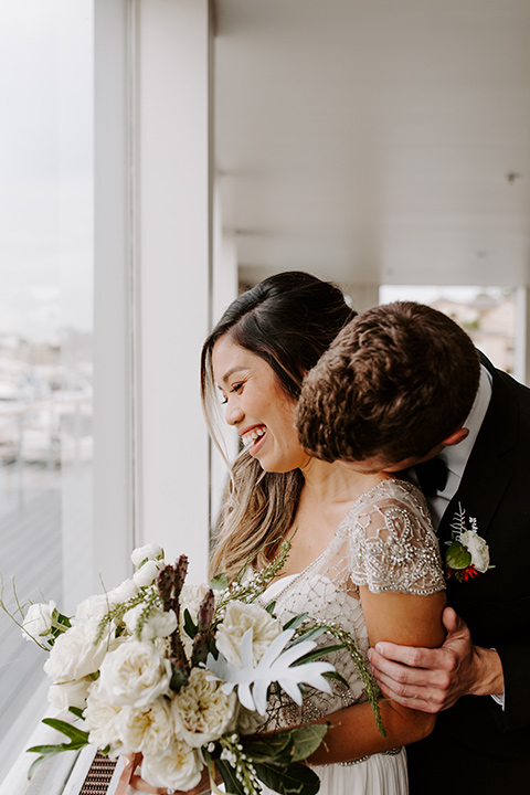 huntington-bay-club-shoot-bride-and-groom-by-window-laughing-bride-wearing-a-flowing-dress-with-a-beaded-bodice-and-sleeves-groom-wearing-a-black-suit-with-a-green-velvet-bow-tie