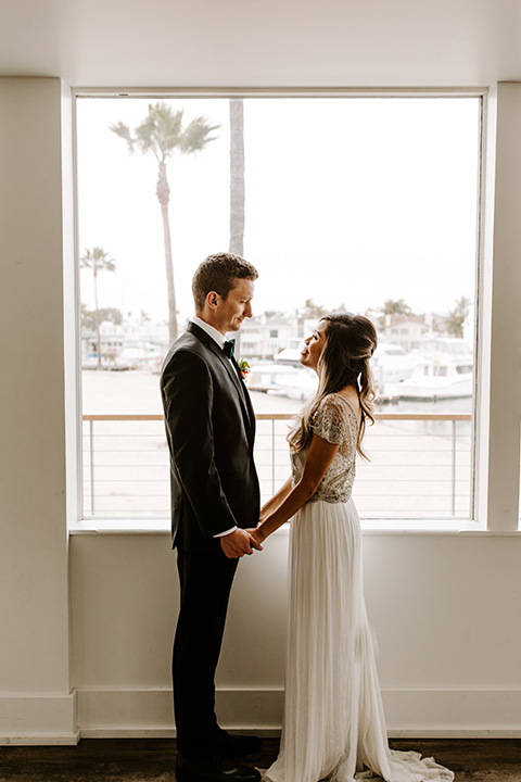 huntington-bay-club-shoot-bride-and-groom-by-windows-wearing-a-flowing-dress-with-a-beaded-bodice-and-sleeves-groom-wearing-a-black-suit-with-a-green-velvet-bow-tie