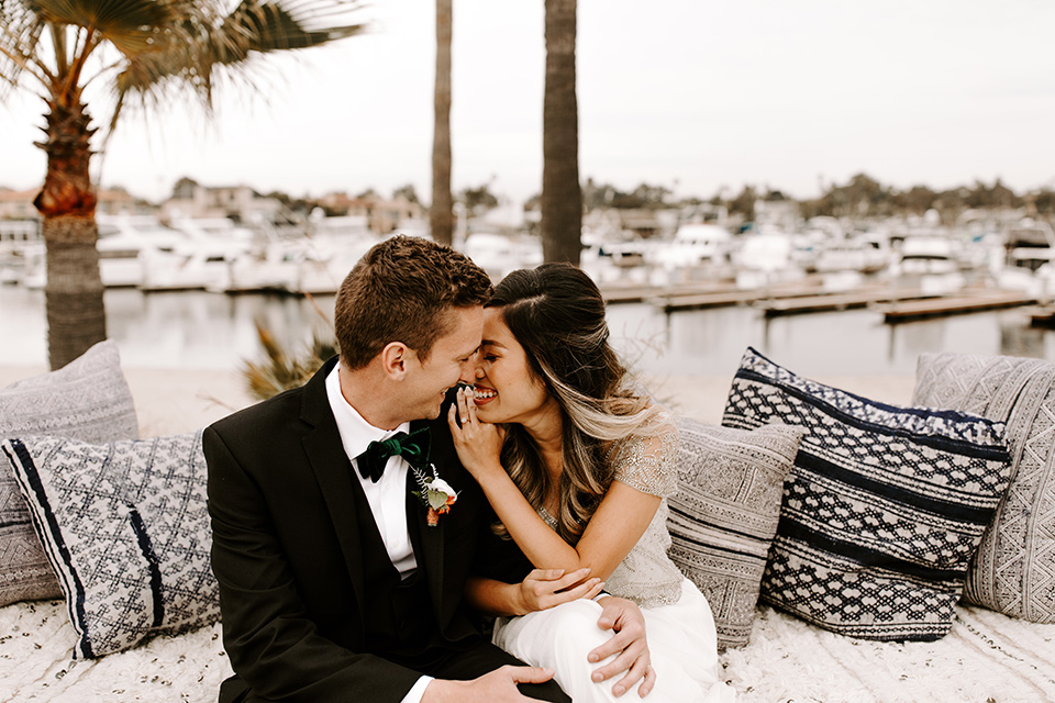 huntington-bay-club-shoot-bride-and-groom-laughing-on-the-couch-bride-wearing-a-flowing-gown-with-a-beaded-bodice-and-ca-sleeves-groom-wearing-a-black-suit-with-a-green-velvet-bow-tie