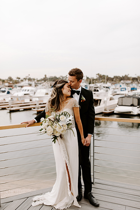 huntington-bay-club-shoot-bride-and-groom-near-the-water-standing-bride-wearing-a-flowing-dress-with-a-beaded-bodice-and-sleeves-groom-wearing-a-black-suit-with-a-green-velvet-bow-tie