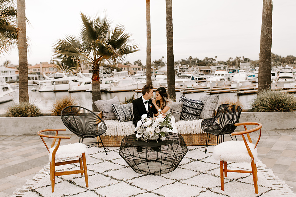 huntington-bay-club-shoot-bride-and-groom-on-couch-bride-wearing-a-flowing-gown-with-a-beaded-bodice-and-ca-sleeves-groom-wearing-a-black-suit-with-a-green-velvet-bow-tie