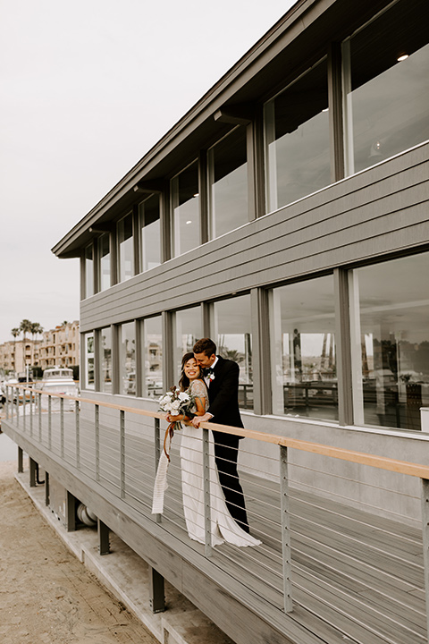 huntington-bay-club-shoot-bride-and-groom-outside-by-metal-awning-bride-wearing-a-flowing-dress-with-a-beaded-bodice-and-sleeves-groom-wearing-a-black-suit-with-a-green-velvet-bow-tie