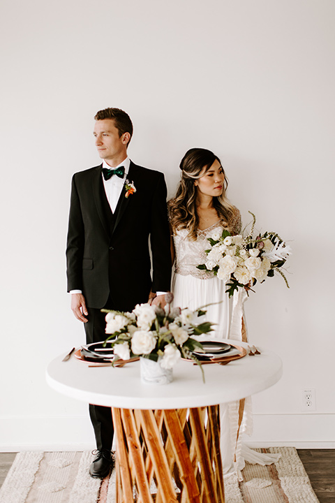 huntington-bay-club-shoot-bride-and-groom-standing-by-table-bride-wearing-a-flowing-dress-with-a-beaded-bodice-and-sleeves-groom-wearing-a-black-suit-with-a-green-velvet-bow-tie