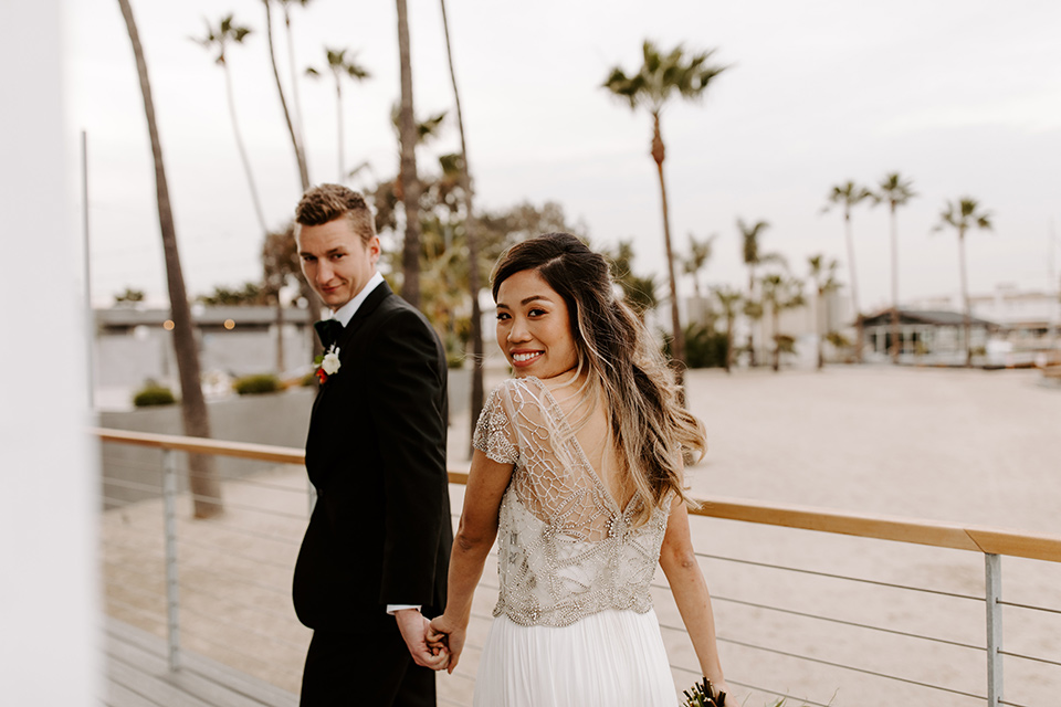 huntington-bay-club-shoot-bride-and-groom-walking-away-looking-over-their-shoulders-bride-wearing-a-flowing-gown-with-a-beaded-bodice-and-ca-sleeves-groom-wearing-a-black-suit-with-a-green-velvet-bow-tie