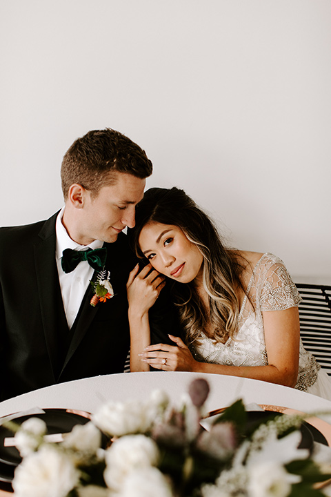 huntington-bay-club-shoot-brode-looking-at-camera-groom-looking-at-her-sitting-bride-wearing-a-flowing-dress-with-a-beaded-bodice-and-sleeves-groom-wearing-a-black-suit-with-a-green-velvet-bow-tie