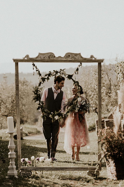 la-dolce-vita-bride-and-groom-at-ceremony-bride-wearing-a-pink-colored-dress-with-lace-detailing-groom-in-grey-pants-and-a-grey-vest