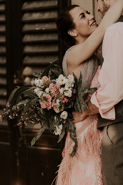 la-dolce-vita-bride-and-groom-holding-each-other-laughing-bride-wearing-a-pink-colored-dress-with-lace-detailing-groom-in-grey-pants-and-a-grey-vest