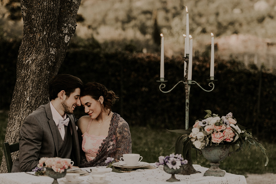 la-dolce-vita-shoot-bride-and-groom-at-sweetheart-table-bride-in-a-pink-colored-dress-with-lace-detailing-groom-in-grey-suit-pants-and-vest