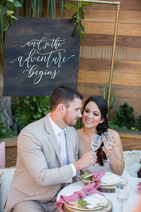 lot-8-bride-and-groom-cheers-sitting-on-pillows-bride-wearing-a-white-strapless-bridal-gown-with-lace-embellishments-hair-in-a-loose-braid-groom-wearing-a-light-grey-suit-with-a-silver-tie