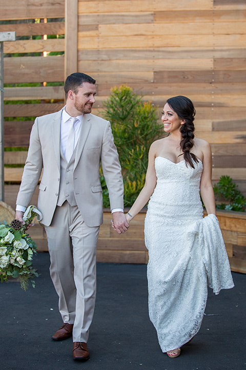 lot-8-bride-and-groom-walking-holding-hands-bride-wearing-a-white-strapless-bridal-gown-with-lace-embellishments-hair-in-a-loose-braid-groom-wearing-a-light-grey-suit-with-a-silver-tie-and-light-brown-shoes