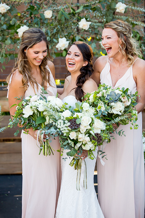 lot-8-bride-with-maids- bride-wearing-a-strapless-white-gown-with-and-loose-braid-while-braideamaids-are-in-blush-dresses-holding-floral-bouquets-and-all-laughing