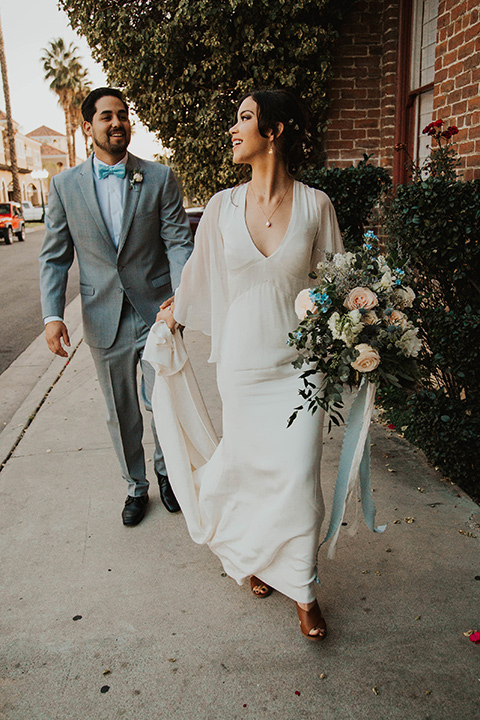 The-mitten-building-shoot-bride-adn-groom-walking-away-outside-bride-in-a-retro-inspired-gown-with-hair-in-a-bun-and-fingerwaves-groom-in-a-light-grey-suit-with-a-blue-bow-tie-and-brown-shoes