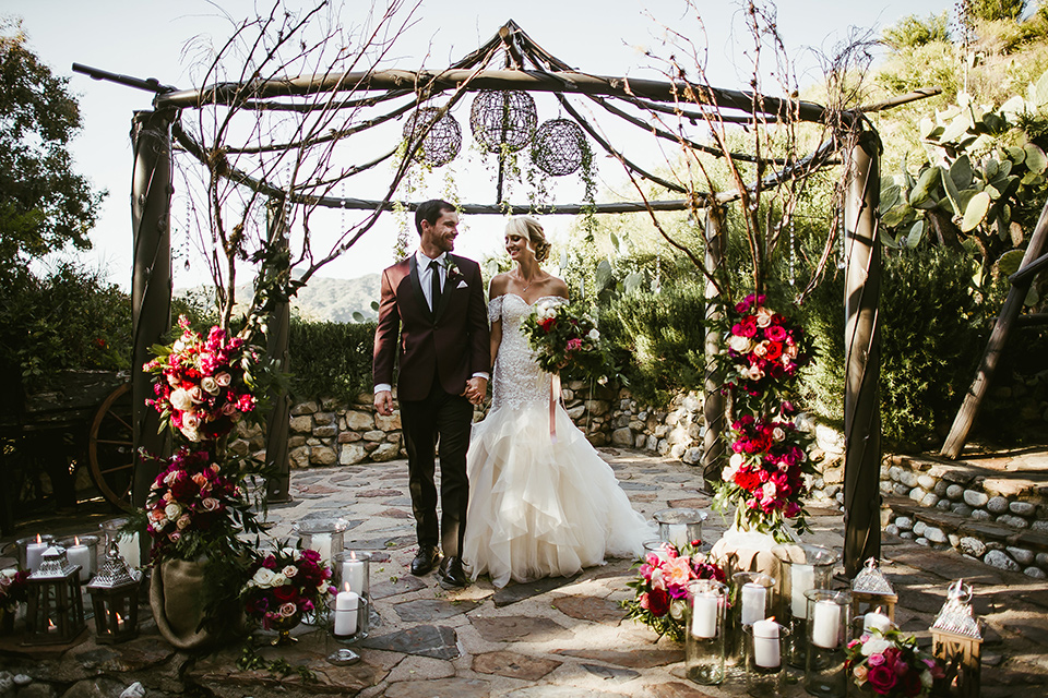 saddle-peak-bride-and-groom-at-ceremony-space-bride-in-an-off-the-shoulder-gown-with-lace-detailing-and-hair-up-in-a-bun-groom-in-a-burgundy-tuxedo-with-a-long-tie