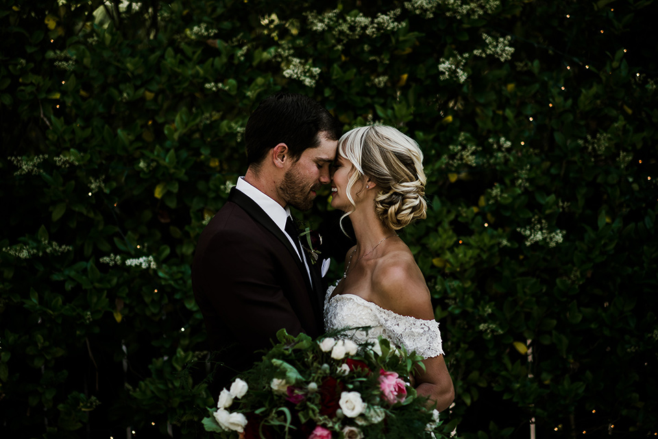 saddle-peak-bride-and-groom-at-ceremony-space-bride-in-an-off-the-shoulder-gown-with-lace-detailing-and-hair-up-in-a-bun-groom-in-a-burgundy-tuxedo-with-a-long-tie