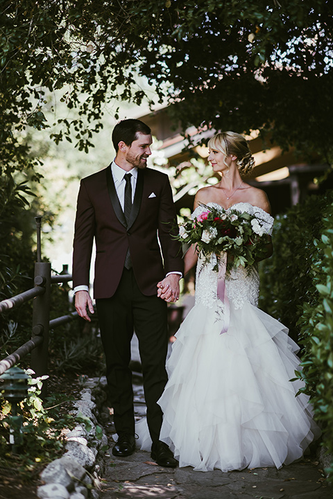 saddle-peak-bride-and-groom-looking-at-eachother-and-walking-bride-in-an-off-the-shoulder-gown-with-lace-detailing-and-hair-up-in-a-bun-groom-in-a-burgundy-tuxedo-with-a-long-tie