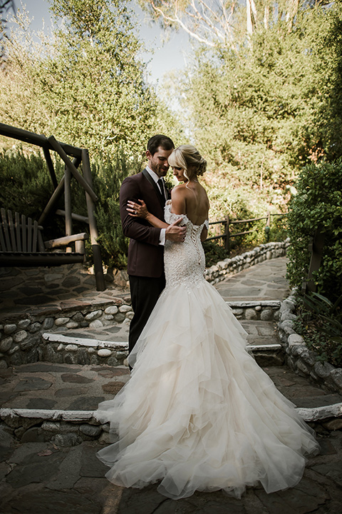 saddle-peak-bride-and-groom-on-steps-bride-in-an-off-the-shoulder-gown-with-lace-detailing-and-hair-up-in-a-bun-groom-in-a-burgundy-tuxedo-with-a-long-tie