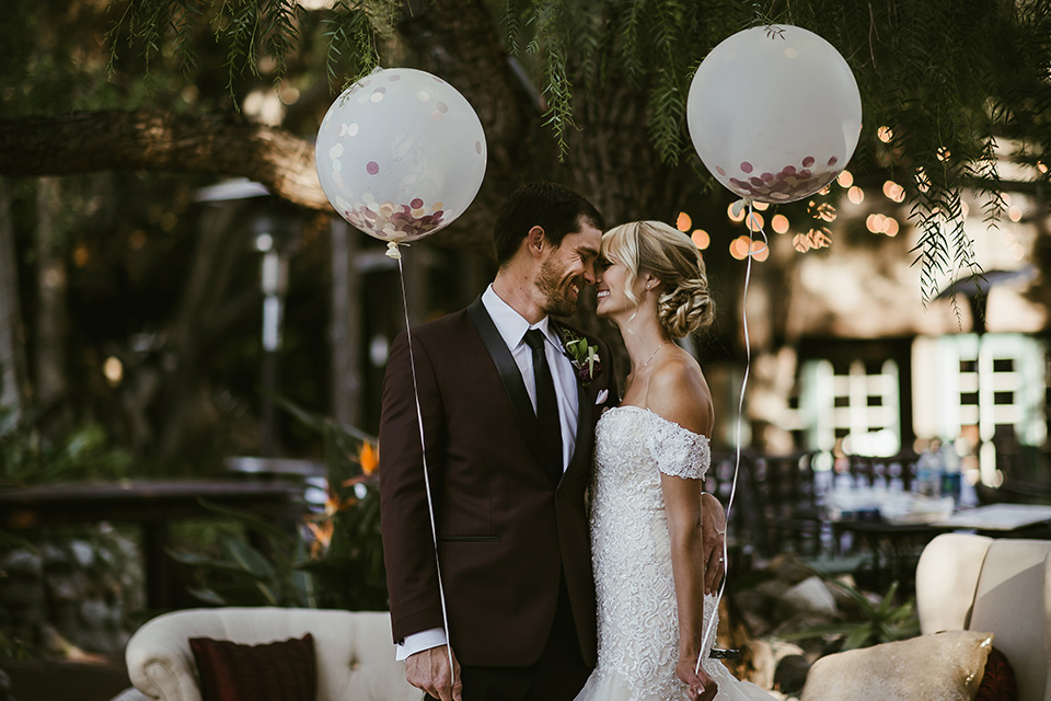 saddle-peak-bride-and-groom-with-balloons-bride-in-an-off-the-shoulder-gown-with-lace-detailing-and-hair-up-in-a-bun-groom-in-a-burgundy-tuxedo-with-a-long-tie