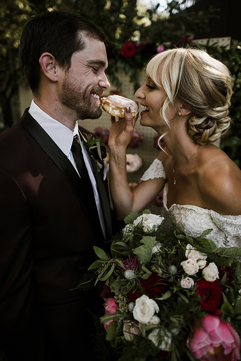 saddle-peak-bride-and-groom-with-donuts-bride-in-an-off-the-shoulder-gown-with-lace-detailing-and-hair-up-in-a-bun-groom-in-a-burgundy-tuxedo-with-a-long-tie