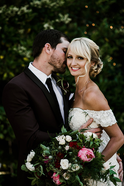 saddle-peak-bride-looking-at-camera-groom-looking-at-her-bride-in-an-off-the-shoulder-gown-with-lace-detailing-and-hair-up-in-a-bun-groom-in-a-burgundy-tuxedo-with-a-long-tie