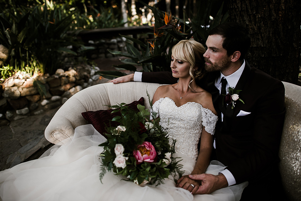 saddle-peak-couple-on-couch-bride-in-an-off-the-shoulder-gown-with-lace-detailing-and-hair-up-in-a-bun-groom-in-a-burgundy-tuxedo-with-a-long-tie