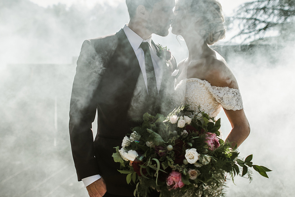 saddle-peak-shoot-bride-and-groom-close-up-in-smoke-bride-in-an-off-the-shoulder-gown-with-lace-detailing-and-hair-up-in-a-bun-groom-in-a-burgundy-tuxedo-with-a-long-tie